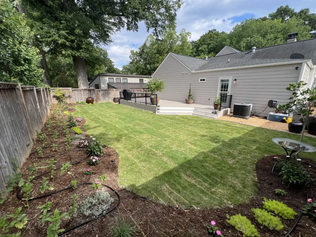 Freshly sodded backyard with mulched beds, drip irrigation along a wood fence, and a small deck behind a gray house.