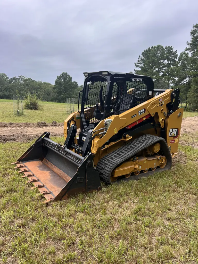 Compact track loader with toothed bucket parked on a grassy field near trees.