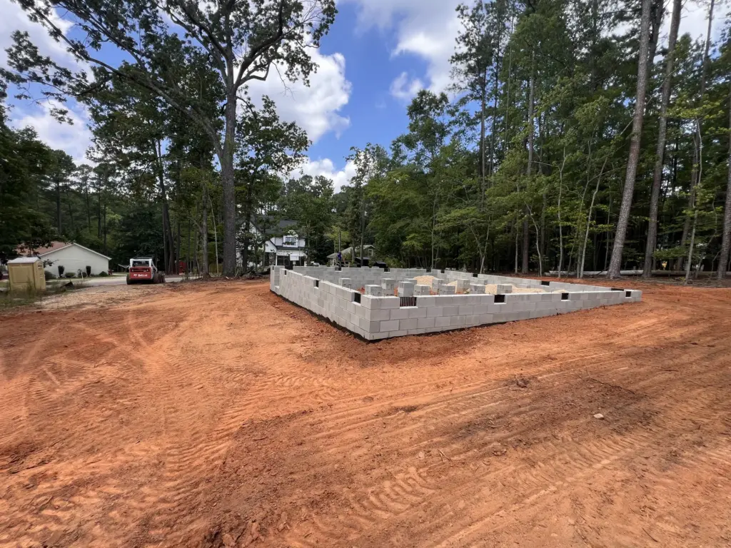 Concrete block foundation on a freshly graded red-clay homesite, surrounded by trees, with equipment in the distance.