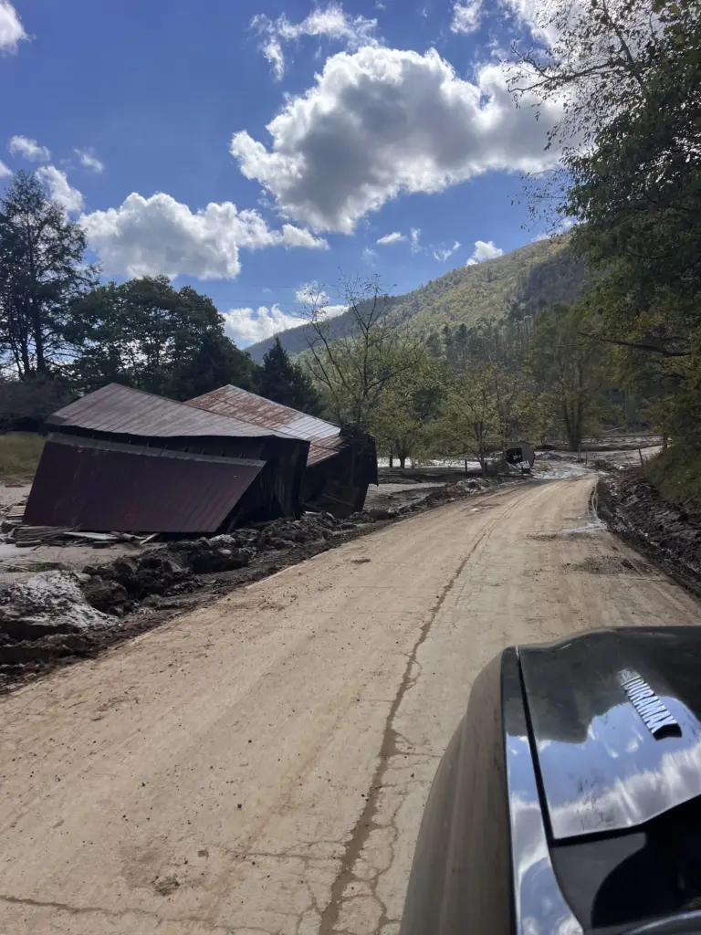 Collapsed barn by muddy rural road after flooding, mountains behind, vehicle hood in foreground.