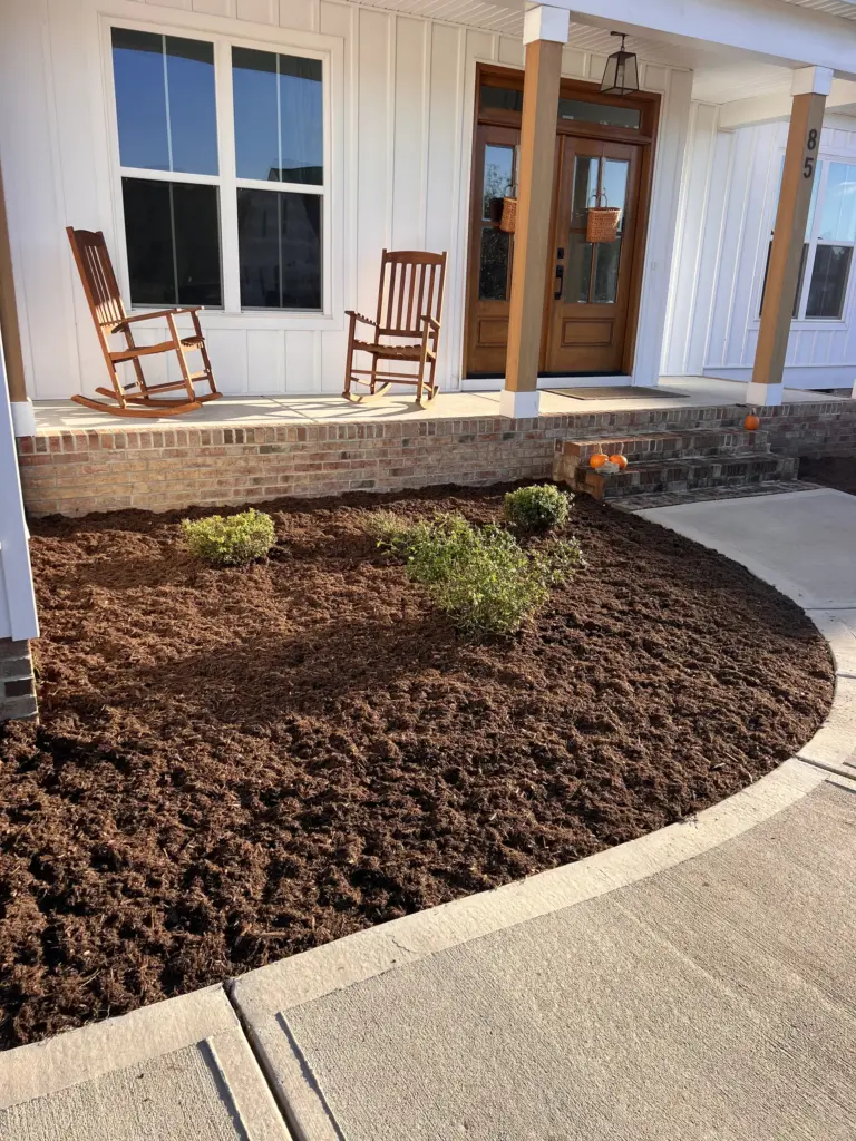 Curved front porch bed freshly mulched with small shrubs beside a concrete walkway and brick steps.