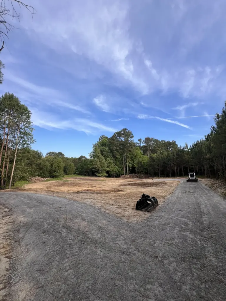 Graded clearing with curved gravel drive, skid steer at right, and coiled drainage pipe, bordered by pines under blue sky.