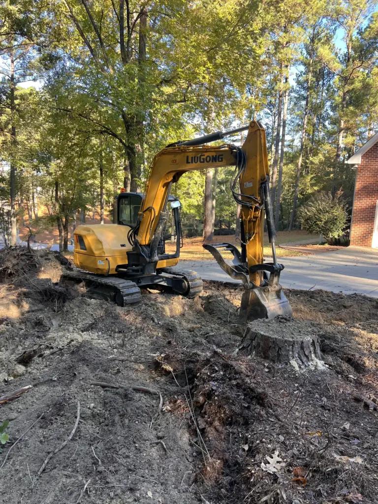 Yellow excavator digging around a tree stump beside a residential driveway under tall trees.