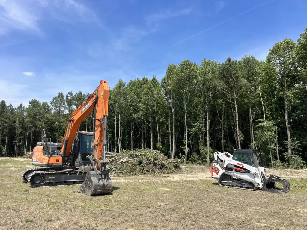 Excavator and compact track loader on a cleared lot beside a brush pile and tree line.