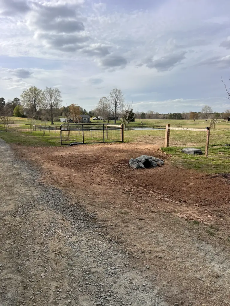 Graded farm entrance with driveway culvert lined with riprap, metal gate and fence, and a gravel lane at left.