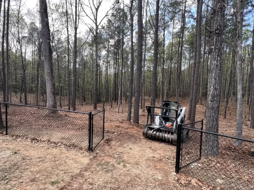 Compact track loader with forestry mulcher behind an open chain-link gate in a wooded backyard.