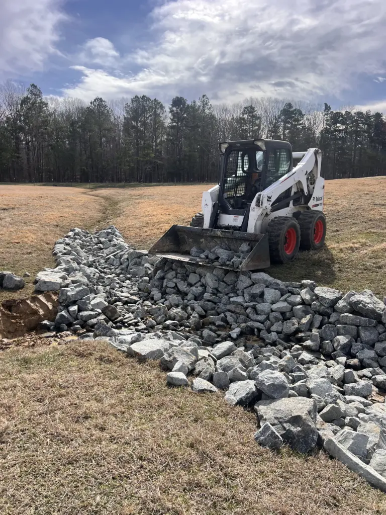 Skid steer spreading riprap rock in a grassy field drainage swale.