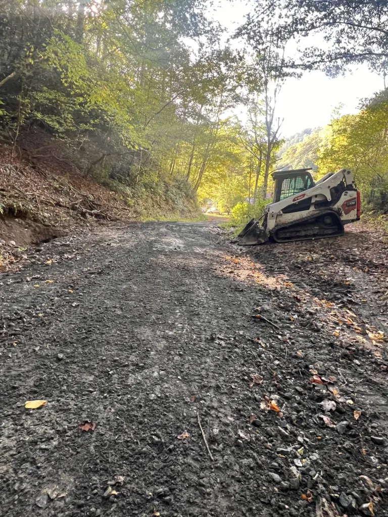 Skid steer parked by a newly graded gravel lane through a wooded hillside.