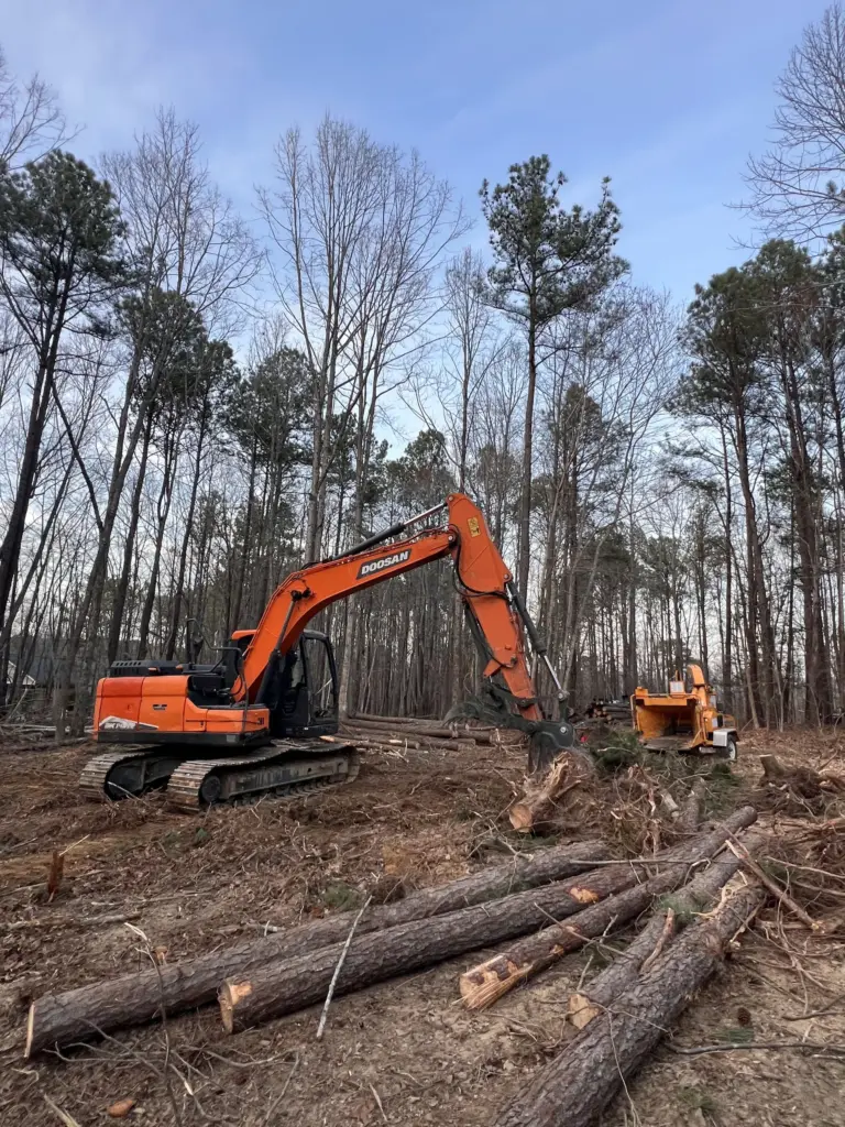 Excavator with grapple stacks cut logs beside a towable wood chipper on a wooded lot clearing.
