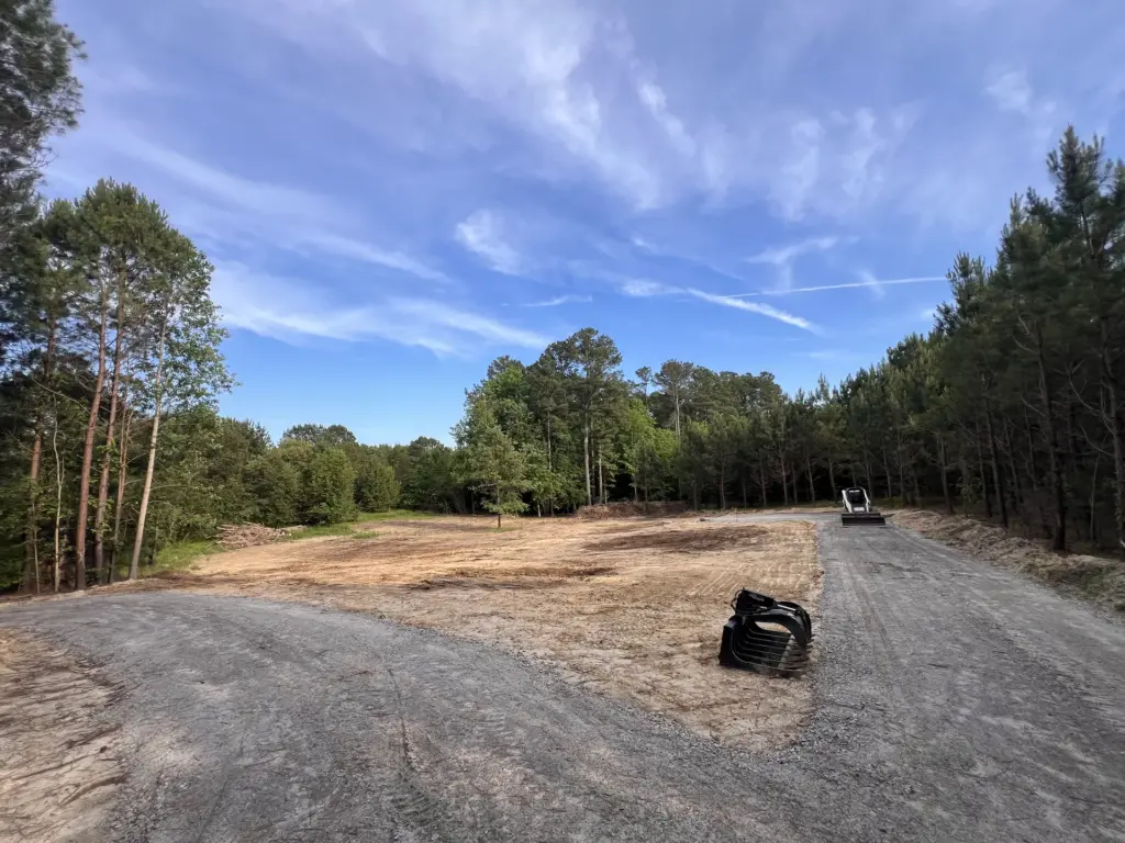 Cleared homesite with gravel driveway through wooded lot, skid steer parked in distance and bucket attachments on the side.