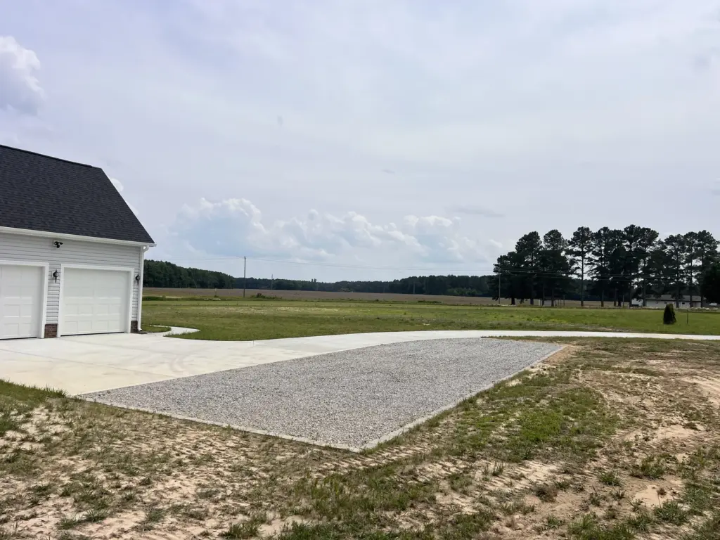 Gravel parking pad beside a concrete driveway at a two-car garage on a rural lot.