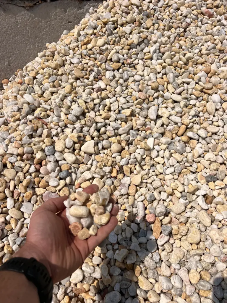 Hand holding tan and white river rock above a large gravel pile outdoors.