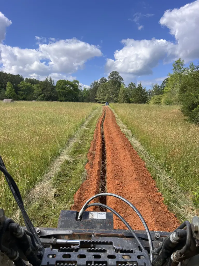 View from a skid steer trencher cutting a long red clay utility trench through a grassy field.