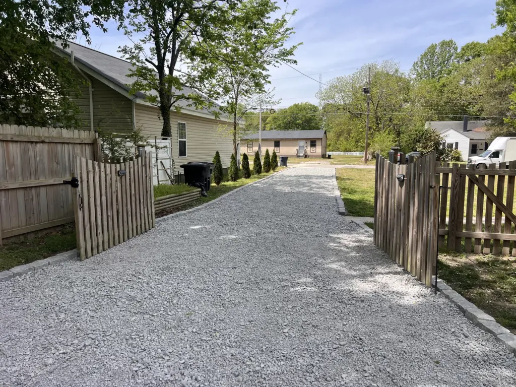 Open wooden gates frame a newly graded gravel driveway beside a house, lined with young evergreens.