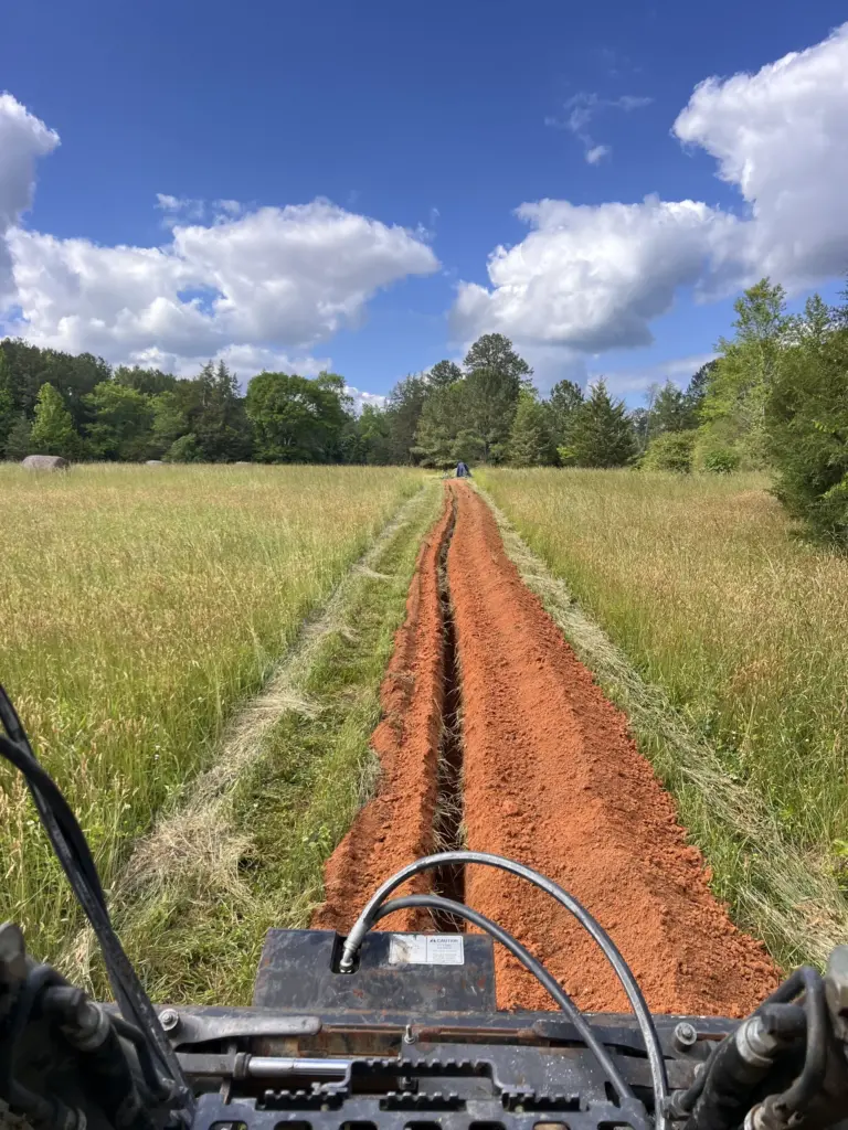 Fresh trench cut through a grassy field by a skid-steer trencher, red clay spoils mounded on both sides.