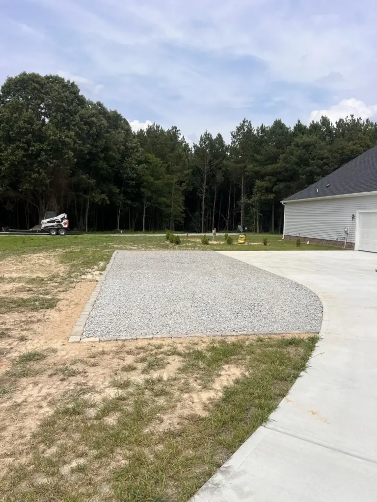 Paver-edged gravel parking pad beside a concrete driveway at a new home, with woods and a skid steer in the background.