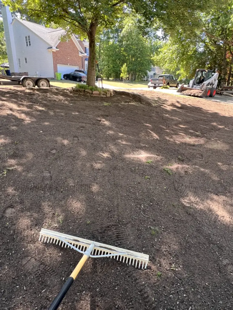 Landscape rake on freshly graded front yard under a tree, with skid steer and trailer by driveway.