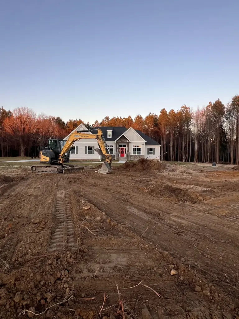Compact excavator grading soil in the front yard of a house with track marks across the lot.