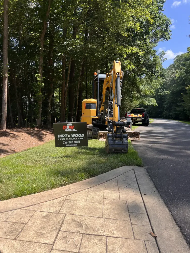 Mini excavator parked by wooded street with yard sign and work truck near a stamped-concrete sidewalk.