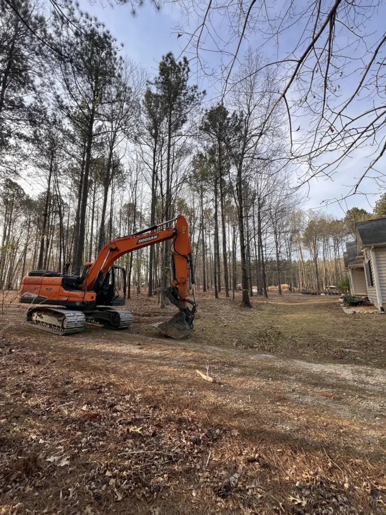 Orange excavator parked on a wooded residential lot beside a house and gravel path.