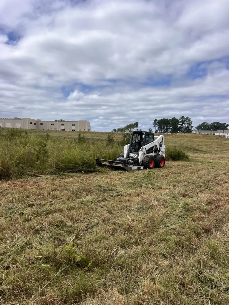 Skid steer with brush cutter mowing tall grass on an industrial lot near warehouses.