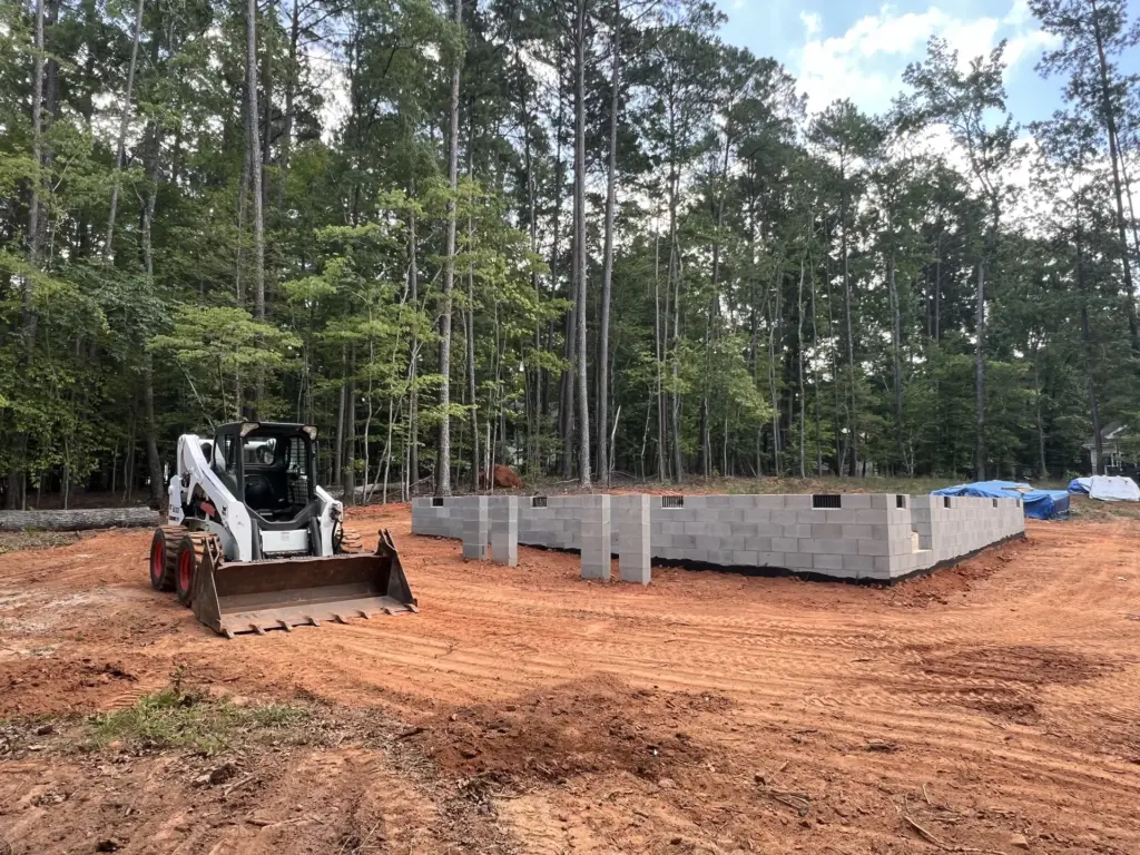 Wheeled skid steer beside a newly built CMU block foundation on a graded red clay lot at the edge of a wooded area.