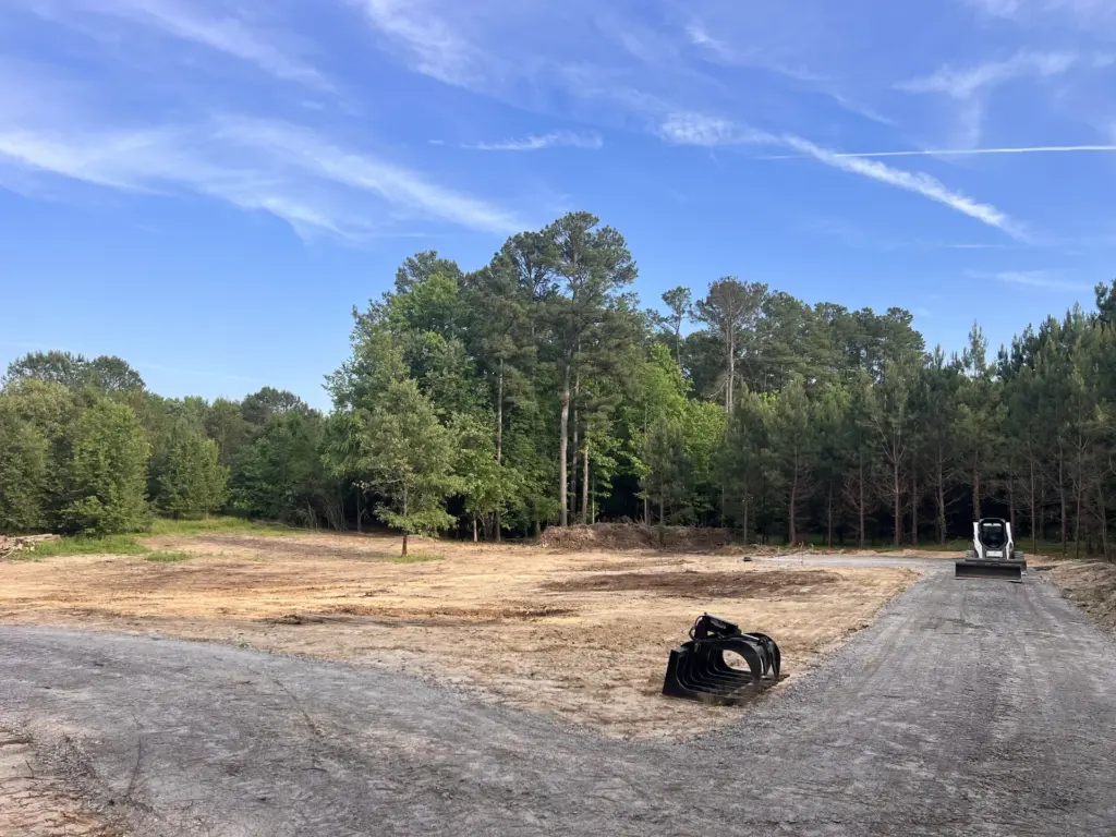 Graded dirt clearing with curved gravel drive, skid steer at right near pine treeline.
