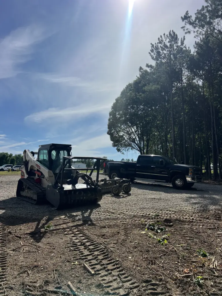 Tracked skid steer with forestry mulcher beside black pickup on a gravel lot near trees.