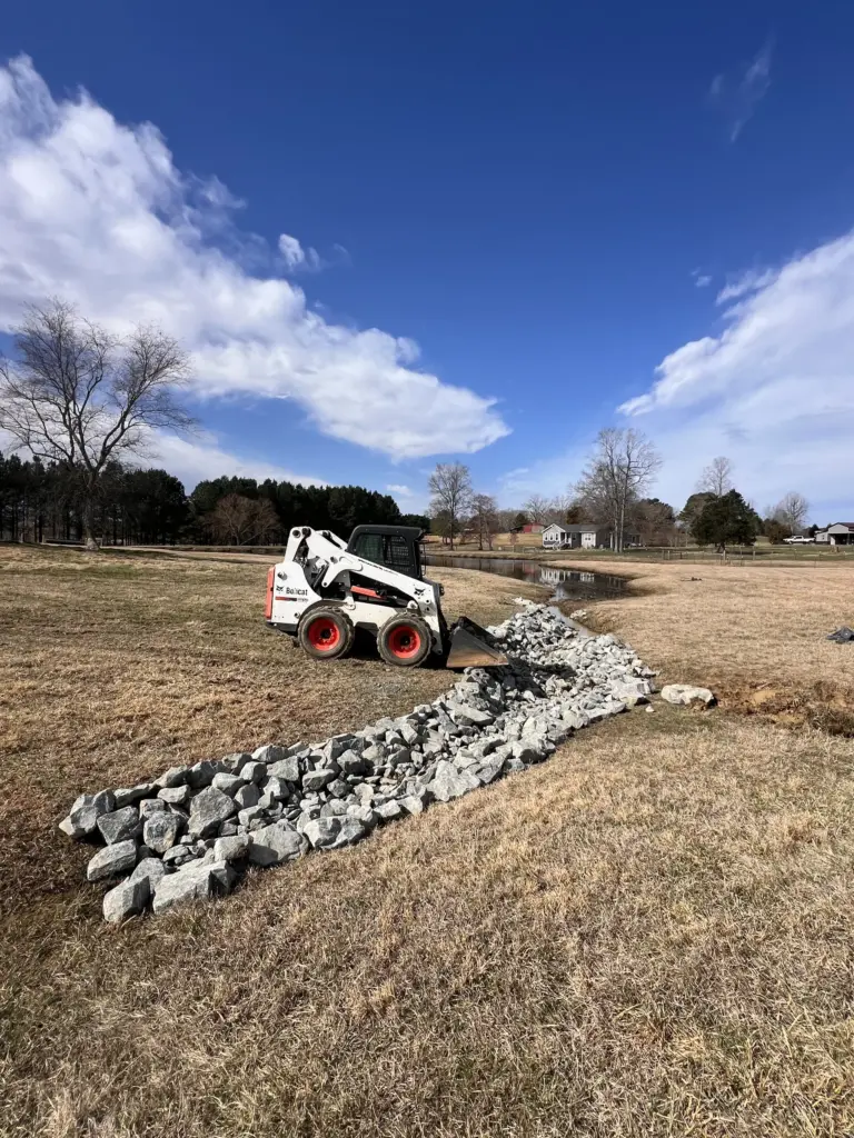 Skid steer spreading riprap rock along a drainage swale in a rural field near a pond.