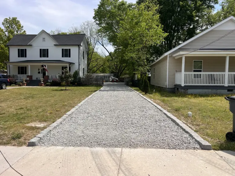 Long crushed-stone driveway with stone edging between two houses, viewed from the street.