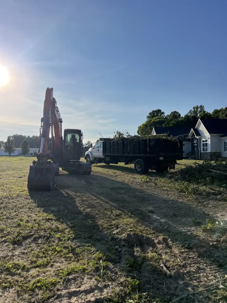 Excavator and dump truck on a cleared residential lot beside houses under a clear sky.