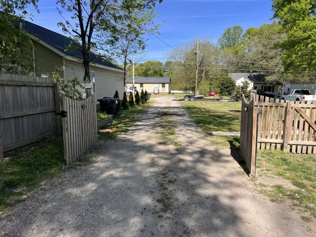 Open wooden gates leading to a narrow gravel driveway between a house and yard.