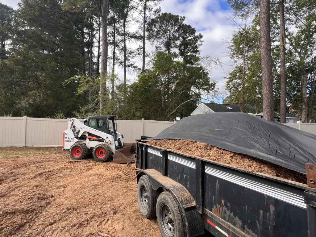Skid-steer loader beside a tarped dump trailer filled with mulch in a fenced backyard.