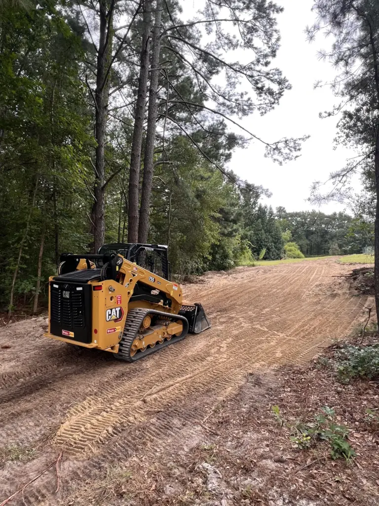 Compact track loader on a freshly graded dirt lane in a wooded area, preparing a path or driveway.