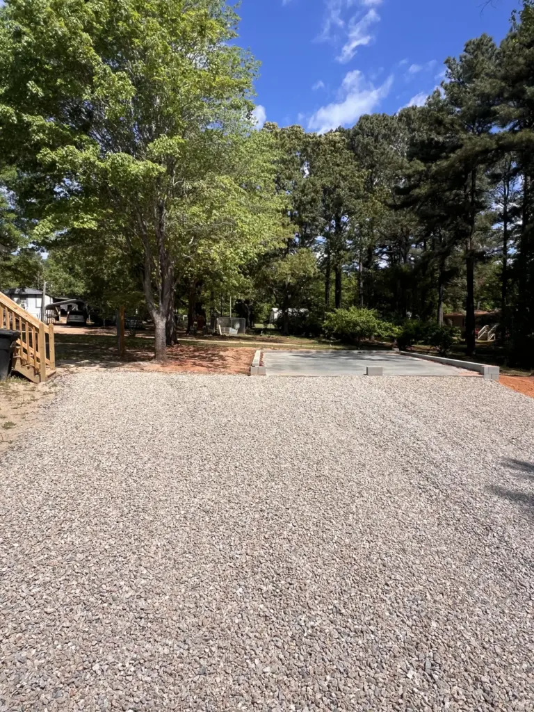 Gravel driveway leading to a new concrete slab in a shaded residential yard with mature trees.