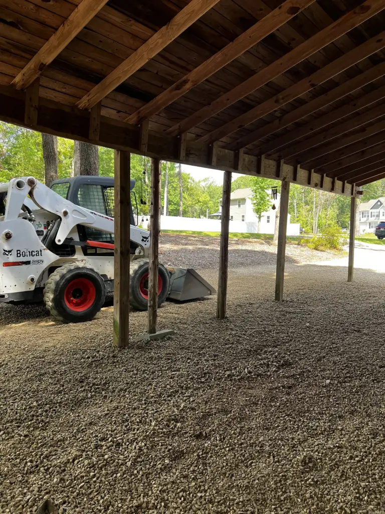 Gravel spread under a wooden carport with a skid-steer parked nearby.