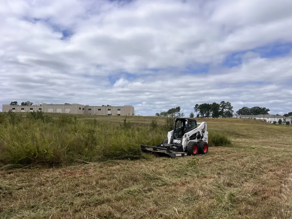 Skid steer with brush cutter mowing an overgrown field near a warehouse complex under cloudy sky in Gallatin, Tennessee.