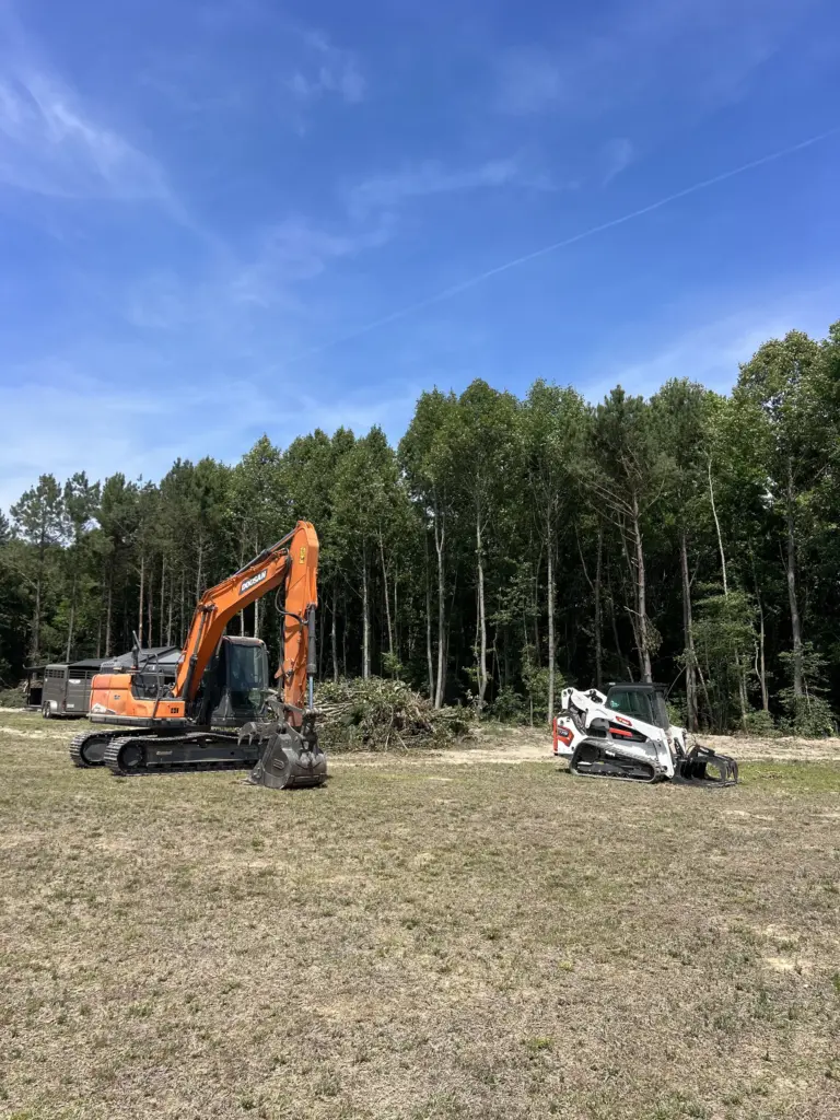 Orange excavator and white skid-steer with grapple parked on a cleared lot beside a tree line under blue sky.