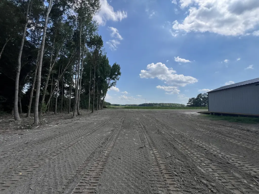 Freshly graded dirt lot with track marks beside a metal shed, tree line on the left, open field under blue sky.