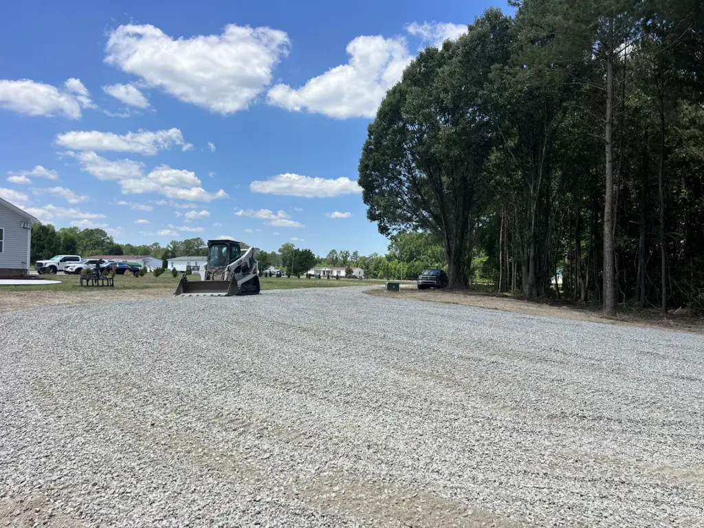 Skid steer parked on a freshly spread gravel parking pad beside trees on a sunny day.