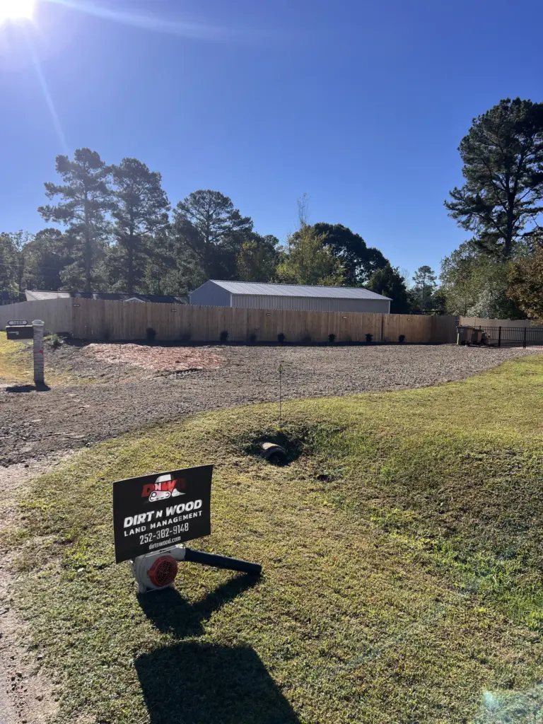 Yard sign on grass in front of a gravel lot, wood privacy fence, and metal-roofed outbuilding under trees.