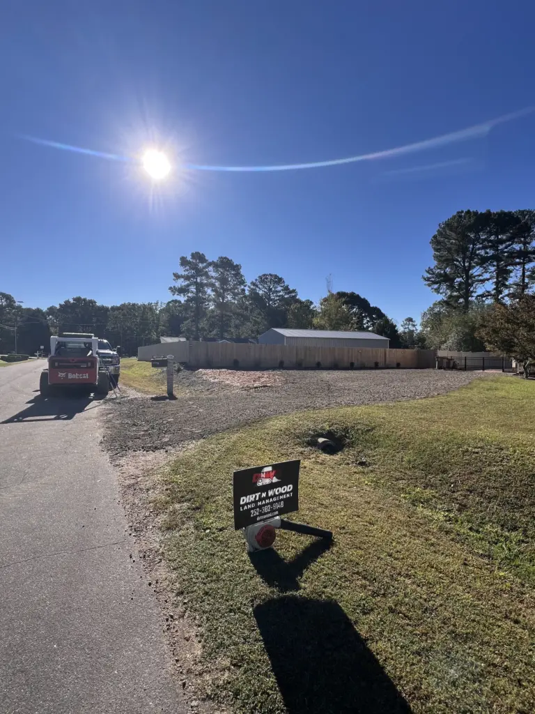 Yard sign by a graded gravel lot and privacy fence on a sunny residential street, with a truck and Bobcat parked at the curb.